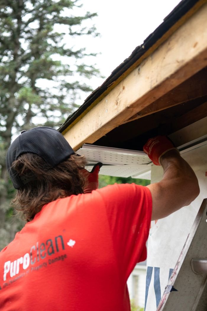 Home A construction worker in a red shirt installs a gutter under a wooden roof, focusing on precision.