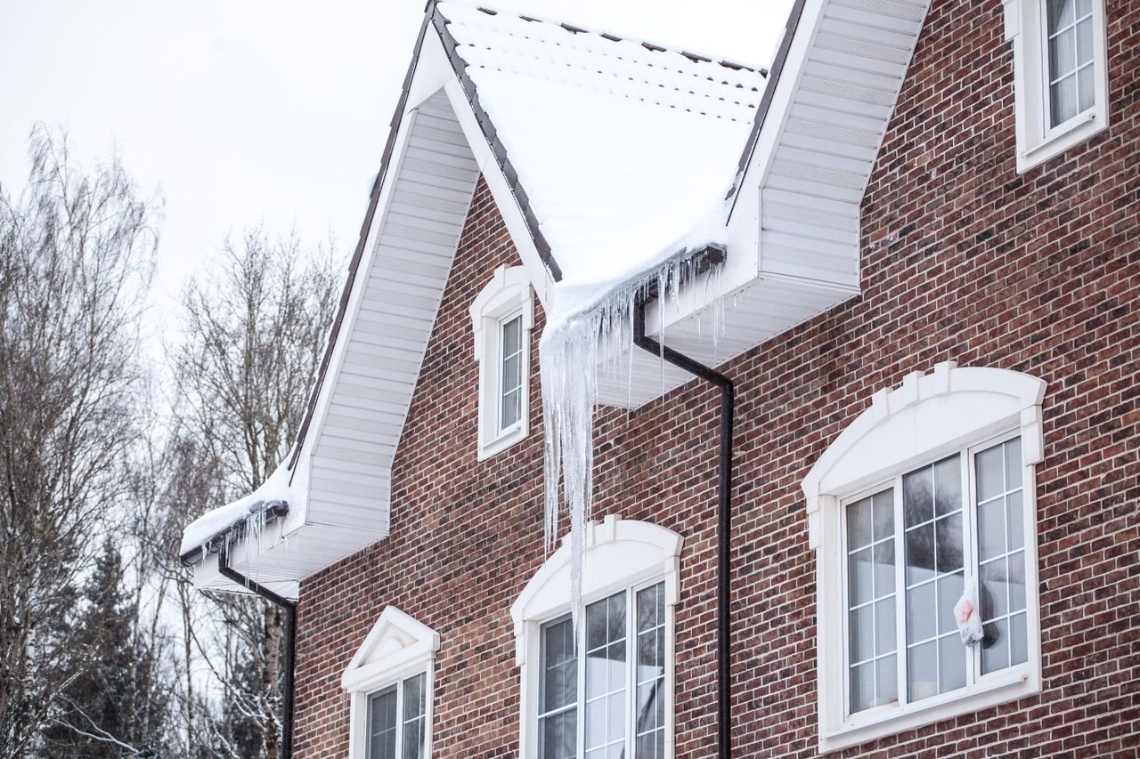 Home A brick house with snow-covered roof and icicles in a winter setting.