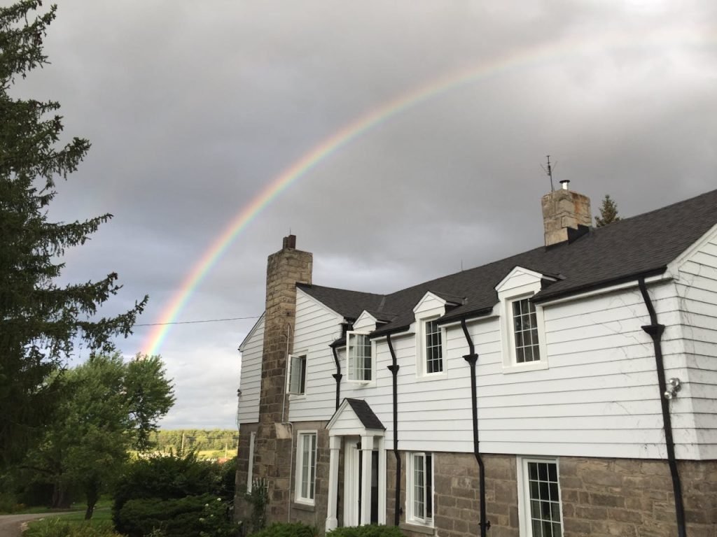 Modern cottage with stone chimney under gloomy sky with glowing colorful rainbow after rain